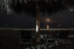 leeg stoelen en rieten parasol Aan zanderig strand Bij nacht foto