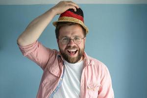 vrolijk Mens in een klein veelkleurig rietje hoed, verhogen het, groeten iemand en glimlachen blij. blauw achtergrond. foto