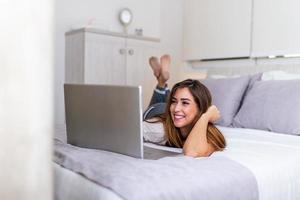 mooi vrouw ontspannende Aan bed gebruik makend van laptop computer. mooi vrouw werken Aan een laptop met glimlachen en aan het liegen naar beneden Aan de bed Bij een condominium in de ochtend. freelancer meisje werken van huis foto