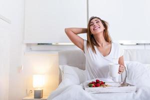 aantrekkelijk vrouw in pyjama ontwaken omhoog in de ochtend- met de geur van koffie en vers ontbijt Aan houten dienblad in bed foto