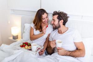 schattig paar hebben ontbijt in bed in de slaapkamer. mooi vrouw voeden haar vriendje aardbeien in bed terwijl hebben ontbijt en koffie in slaapkamer foto