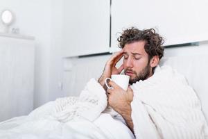 portret van een ziek Mens met de griep, allergie, ziektekiemen, koude hoesten. ziek Mens met hoofdpijn zittend onder de deken met hoog koorts en een griep, resting en drinken heet drank foto