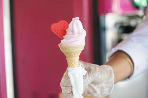 een arbeider met handschoenen Holding een ijs room ijshoorntje met een mengen smaken roze en vanille. roze ijs room met een rood hart. foto