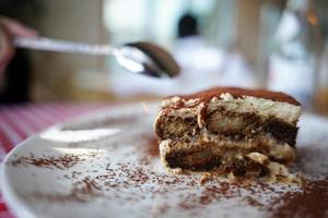 een persoon is aan het eten een tiramisu Italiaans toetje bekroond met koffie poeder en geserveerd Aan een bord Bij een luxe rood cheques patroon tafel met een voorkant visie. foto