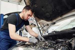 onder de kap. Mens in werk uniform reparaties wit auto- binnenshuis. opvatting van auto- onderhoud foto