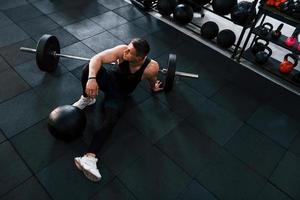 sterk Mens in sportief kleren zittend Aan de verdieping in de Sportschool met uitrusting foto