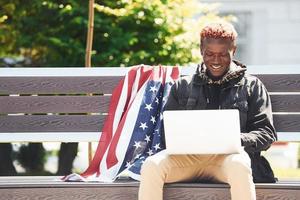 Verenigde Staten van Amerika vlag leunend achter. gebruik makend van laptop. jong Afrikaanse Amerikaans Mens in zwart jasje buitenshuis in de stad zittend Aan bank foto