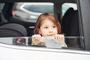 schattig weinig meisje looks door venster auto. opvatting van op reis en vakantie foto