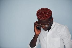 jong Afrikaanse Amerikaans Mens in wit formeel kleren pratend door de telefoon tegen muur binnenshuis foto