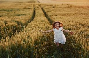 vrolijk weinig meisje in wit jurk rennen in de agrarisch veld- Bij zomer dag tijd foto