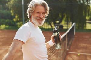 nemen een breken en drinken water. senior modern elegant Mens met racket buitenshuis Aan tennis rechtbank Bij dag foto