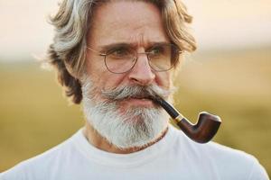 portret van senior elegant Mens met grijs haar- en baard dat staand buitenshuis Aan veld- Bij zonnig dag en roken foto