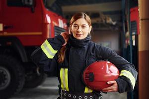 houdt rood hoed in handen. vrouw brandweerman in beschermend uniform staand in de buurt vrachtauto foto