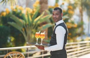 houdt cocktails. zwart ober in formeel kleren is Bij zijn werk buitenshuis Bij zonnig dag foto