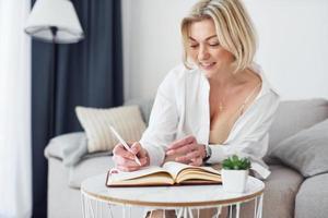 middelbare leeftijd vrouw in elegant kleren is Bij huis schrijven in kladblok foto