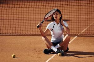 brunette is zittend Aan de grond. vrouw tennis speler is Aan de rechtbank Bij dag foto