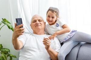 portret van de schattig meisje spelen of typen Aan de smartphone terwijl zittend Aan de bankstel met haar opa Bij huis. foto