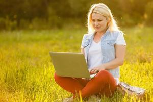 jong vrouw met laptop zit Aan de gras in de park Aan een zonnig dag. lachend blond in een wit tank top en korte broek. online opleiding, afgelegen werk en communicatie in sociaal netwerken foto