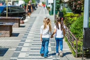 twee school- meisjes vervelend rugzak buiten de primair school. schoolmeisje, elementair school- leerling gaan van school, diploma uitreiking, zomer vakantie. foto