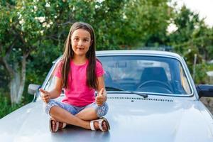 weinig meisje zittend Aan de kap. reparatie oud, gebroken auto. foto