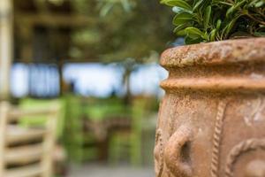 antiek terracotta vaas detailopname, middellandse Zee zomer stemming, buitenshuis restaurant met tropisch bloemen en bladeren, wazig stoelen en tafel. foto