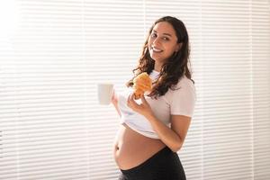 mooi zwanger vrouw Holding croissant en kop van koffie in haar handen gedurende ochtend- ontbijt. concept van mooi zo Gezondheid en positief houding terwijl ervan uitgaand baby foto