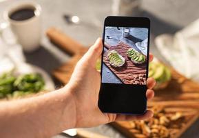 handen nemen afbeeldingen Aan smartphone van twee mooi gezond verzuren room en avocado boterhammen aan het liegen Aan bord Aan de tafel. sociaal media en voedsel concept foto