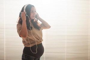 krullend haar brunette gepacificeerd zwanger vrouw luistert naar aangenaam klassiek muziek- gebruik makend van smartphone en koptelefoon. concept van een rustgevend humeur voordat vergadering baby. copyspace foto