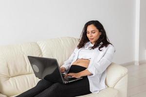 zwanger vrouw gebruik makend van laptop terwijl zittend Aan een bankstel in de leven kamer Bij huis foto