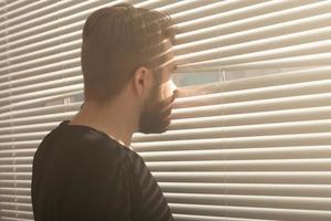 achterzijde visie van jong Mens met baard gluurt door gat in de venster jaloezieën en looks uit in de straat. toezicht en nieuwsgierigheid concept foto