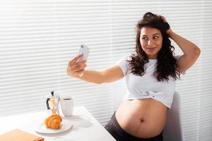 zwanger vrouw duurt selfie Aan haar smartphone terwijl ontbijt. moederschap vertrekken en technologieën concept. foto