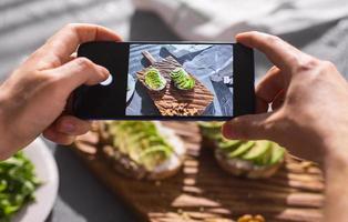 handen nemen afbeeldingen Aan smartphone van twee mooi gezond verzuren room en avocado boterhammen aan het liegen Aan bord Aan de tafel. sociaal media en voedsel concept foto