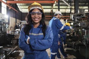 portret van professioneel zwart vrouw arbeider in beschermend veiligheid uniform en helm op zoek Bij camera, armen gekruiste en lachend, met ingenieurs team achter haar in een metaalwerk fabricage fabriek. foto