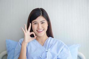 vrouw geduldig, mooi glimlach, Aziatisch zittend in een ziekenhuis bed, duimen omhoog. Oke. de concept van medisch Diensten, Gezondheid verzekering. foto