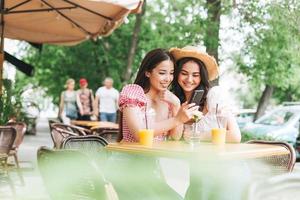 gelukkig glimlachen mooi brunette jong Dames vrienden in zomer kleren gebruik makend van mobiel telefoon in de cafe Bij zomer stad stree foto