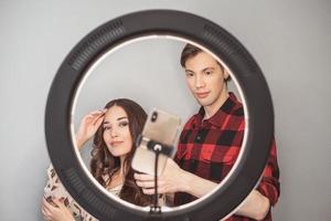 jong paar van schoonheid bloggers, haar- stilist, kapper en model- met haar- stijl schieten Aan mobiel telefoon met ring lamp Aan de grijs muur achtergrond foto