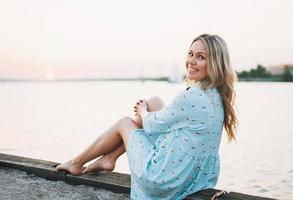 mooi blond jong vrouw in blauw jurk zittend Aan pier en op zoek Aan de zonsondergang foto
