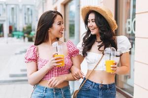 gelukkig glimlachen mooi brunette jong Dames vrienden in zomer kleren met sap in handen wandelen Aan de zomer stad straat foto