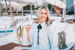 mooi blond jong vrouw in blauw jurk met glas van Frisdrank Aan boot Bij pier in de zonsondergang tijd foto