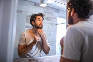 reflectie van knap Mens met baard op zoek Bij spiegel en aanraken gezicht in badkamer poetsen. Mens zetten huidsverzorging gelaats behandeling room Aan gezicht. foto