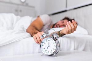 Mens aan het liegen in bed draaien uit een alarm klok in de ochtend- Bij 7 uur. hand- bochten uit de alarm klok ontwaken omhoog Bij ochtend, Mens bochten uit de alarm klok ontwaken omhoog in de ochtend- van een telefoongesprek. foto