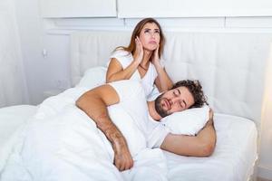 Mens snurken terwijl zijn vrouw is aan het bedekken oren met handen. vrouw lijdt van haar mannetje partner snurken in bed. lawaai concept. echt mensen. jong geërgerd vrouw aan het liegen in bed met snurken man foto