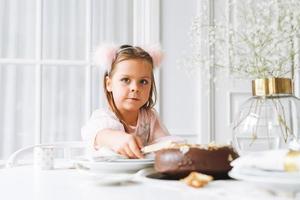 grappig schattig weinig meisje met lang haar- in licht roze jurk met chocola taart in handen Aan feestelijk tafel in helder leven kamer Bij huis. Kerstmis tijd, verjaardag meisje foto