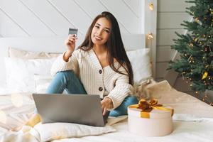 jong mooi Aziatisch vrouw met donker lang haar- in knus wit gebreid trui met Cadeau geschenk doos met gouden lint gebruik makend van laptop met credit kaart Aan bed in kamer met Kerstmis boom Bij huis foto