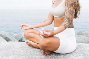 Bijsnijden foto van jong fit mooi vrouw met lang haar- in wit sport kleren praktijk yoga en genieten leven Aan de zee strand, mentaal en fysiek Gezondheid