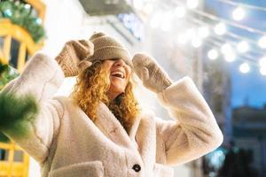 portret van jong gelukkig vrouw met gekruld haar- in vacht jasje hebben pret in winter straat versierd met lichten