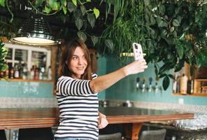 jong aantrekkelijk glimlachen vrouw in gewoontjes kleren gebruik makend van sociaal media Aan mobiel telefoon nemen selfie Bij de cafe foto