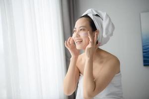 gelukkig jong Aziatisch vrouw toepassen gezicht lotions terwijl vervelend een handdoek en aanraken haar gezicht. dagelijks bedenken en huidsverzorging foto