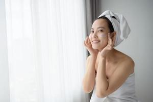 gelukkig jong Aziatisch vrouw toepassen gezicht lotions terwijl vervelend een handdoek en aanraken haar gezicht. dagelijks bedenken en huidsverzorging foto