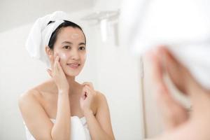 gelukkig jong Aziatisch vrouw toepassen gezicht lotions terwijl vervelend een handdoek en aanraken haar gezicht in badkamer foto